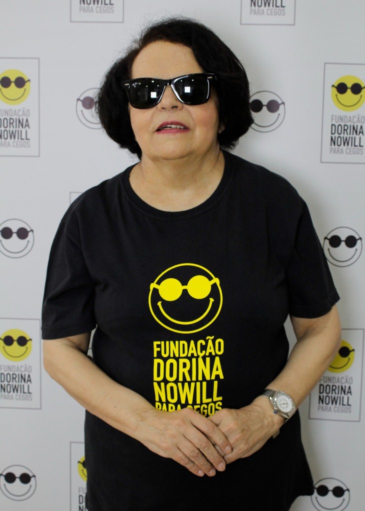 Foto di Regina che veste una t-shirt nera con uno smile giallo e occhiali da sole. Dietro a Regina c’è una parete con tanti smile in occhiali da sole simili a quello della sua maglietta