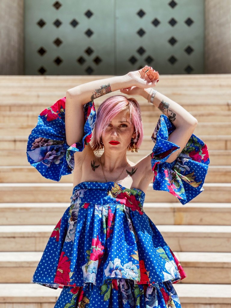 Foto all’aperto di Alexandra. Dietro a lei una scalinata in pietra. Alexandra sfoggia unabito blu della sua produzione. È cosparso di puntini azzurri e di fiori rossi e azzurri. Alexandra ha i capelli rosa  e sorride alla fotocamera
