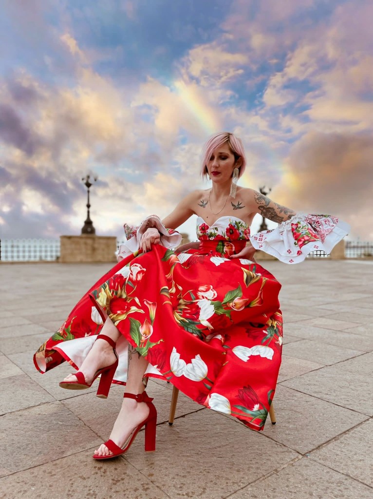 Foto di Alexandra che posa seduta su una piazza. Indossa un abito della sua collezione tutto rosso e composto da gonna e maglietta senza spalline. Ovunque è decorato con fiori bianchi e rossi e foglie. Alexandra ha i capelli rosa e sorride alla fotocamera.