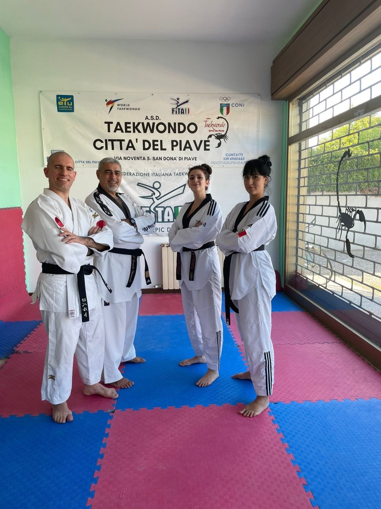 Gruppo in posa a forma di lettera Vu. In piedi sul tatami. Tutti indossano il Dobok a parte Roberto in judogi. Da sinistra, Roberto, Maestro Chiumento, Isabella e Michelle. Sul fondo, l’insegna del Taekwondo Città del Piave.