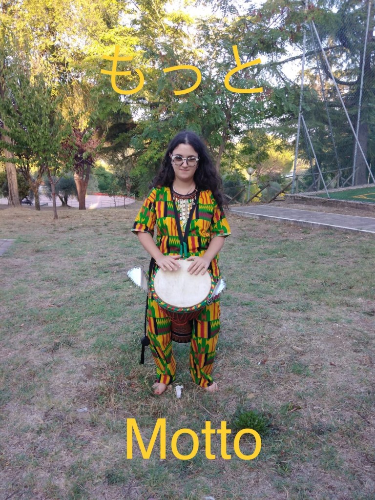 Clarissa seduta nei giardini con in mano uno jambe. Indossa occhiali da sole e una camicia multicolore verde, gialla e rossa