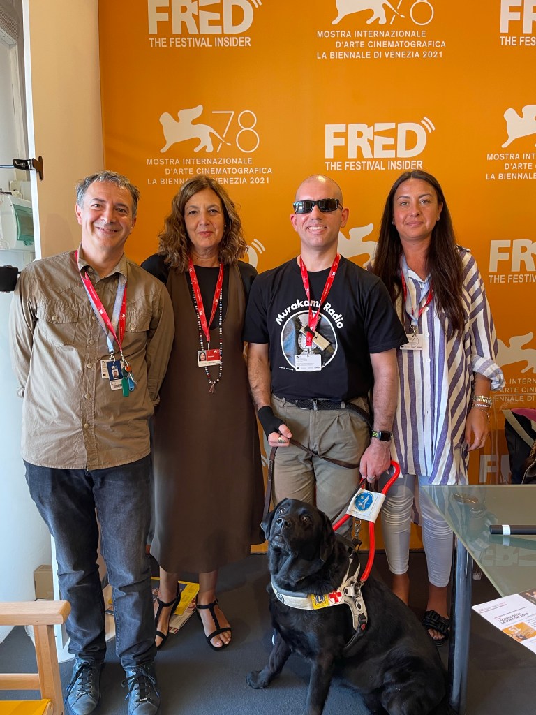 Foto di gruppo a FRED Radio. Sfondo arancione e davanti a tutti Atena, il cane guida di Roberto che guarda verso l’obiettivo

