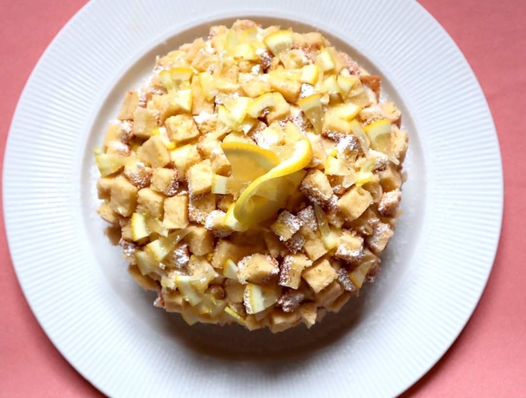 Su un piatto bianco giace la torta Mimosa. La sua forma a cupola è ricoperta da tanti piccoli pezzi di pan di spagna
per ricordare la forma del fiore della mimosa.
Il colore è molto giallo, la torta è guarnita con piccoli pezzi tagliati di limone
e zucchero a velo.