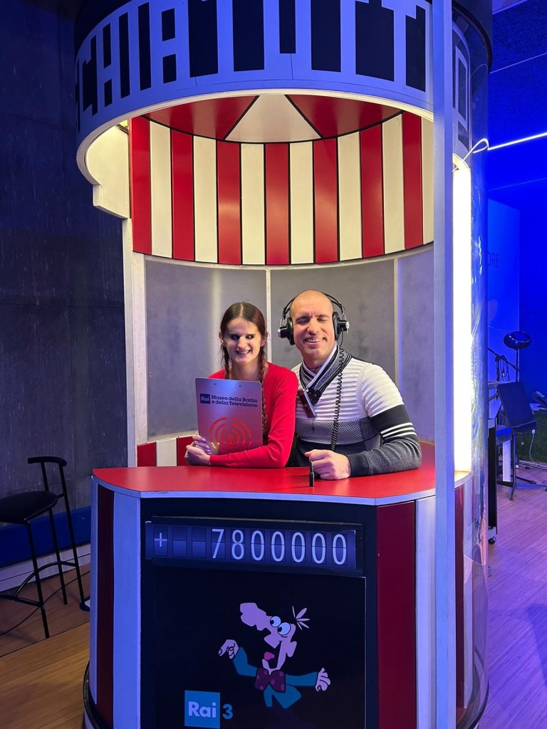 Elena e roberto siedono dentro la cabina di rischiatutto. Elena tiene in mano la cartellina rai da presentatrice, roberto indossa le cuffie da concorrente