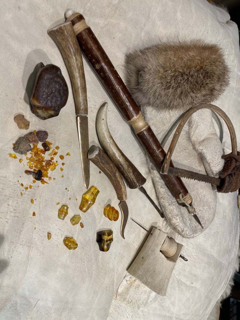 Sopra un velluto bianco  sono adagiati tirinti di diverse misure e scarti di ambra nella parte bassa. Nella parte alta un guanto in pelle che si usava nel baltico e alcuni strumenti per lavorare l’ambra
